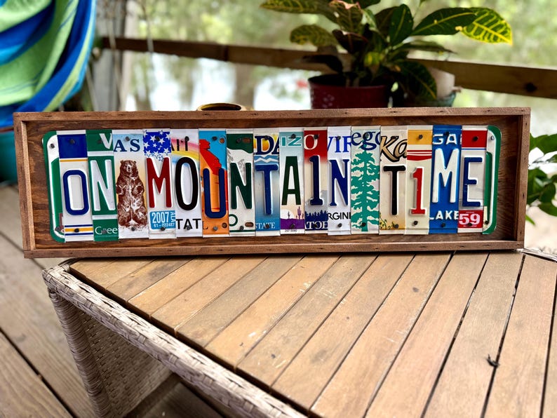May include: A rectangular wooden sign featuring the text ON MOUNTAIN TIME spelled out using various colorful vintage license plates. The plates display diverse state graphics, including a bear, a pine tree, and various letters, all framed within a dark brown wooden border.