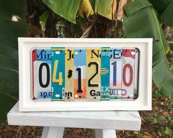 May include: A framed artwork made from vintage license plates, featuring the numbers 04-12-10. The license plates are arranged vertically and are in various colors, including red, blue, yellow, and green.