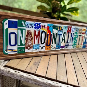 May include: A rectangular wooden frame containing a collage of various colorful vintage license plates arranged to spell out the phrase ON MOUNTAIN TIME. The artwork features a central brown bear graphic and diverse state plate designs.