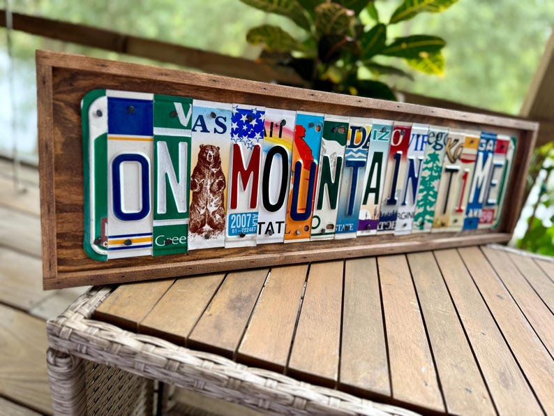 May include: A rectangular wooden frame containing a collage of various colorful vintage license plates arranged to spell out the phrase ON MOUNTAIN TIME. The artwork features a central brown bear graphic and diverse state plate designs.