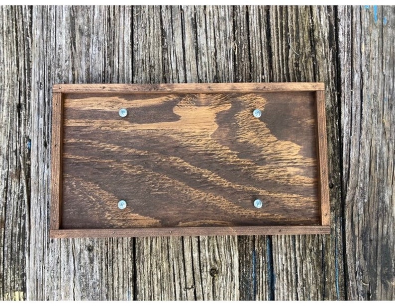 May include: A rectangular wooden sign with a dark brown stain. The sign has four screws holding it together. The sign is on a wooden background.