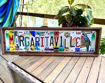 May include: A rectangular wooden frame containing a collage of various colorful license plates arranged to spell out the word MARGARITAVILLE. The letters are formed by segments of different state license plates, featuring a parrot graphic on the right.