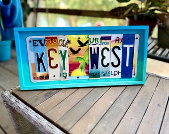 May include: A rectangular wall sign featuring the text KEY WEST spelled out using segments of various colourful license plates. The sign is encased in a bright turquoise wooden frame and includes imagery of palm trees and birds.