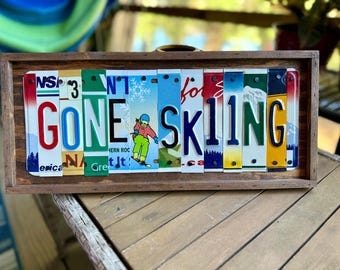 May include: A rectangular wooden sign featuring the text GONE SKIING spelled out using vertical segments of various colorful license plates. The design includes a graphic of a person skiing and is framed in a dark brown wood border.