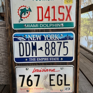 May include: A wooden frame with three license plates. The top plate is from Florida and says "D41SX" with the Miami Dolphins logo. The middle plate is from New York and says "DDM-8875". The bottom plate is from Louisiana and says "767 EGL".