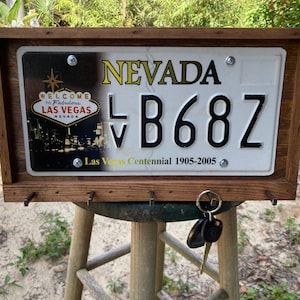 May include: A rectangular wooden frame holding a Nevada license plate with the text Welcome to Fabulous Las Vegas Nevada and Las Vegas Centennial 1905-2005. The plate features the characters LV B68Z and includes four metal hooks for hanging keys.
