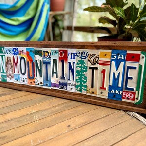 May include: A rectangular wooden frame containing a collage of various colorful license plates arranged to spell out the phrase ON MOUNTAIN TIME. The plates feature diverse state designs, fonts, and graphics, including a pine tree and a bear silhouette.