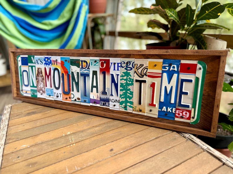 May include: A rectangular wooden frame containing a collage of various colorful license plates arranged to spell out the phrase ON MOUNTAIN TIME. The plates feature diverse state designs, fonts, and graphics, including a pine tree and a bear silhouette.