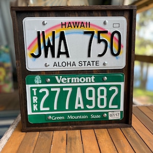 May include: A wooden frame with two license plates inside. The top plate is white with a rainbow and the text "HAWAII JWA 750 ALOHA STATE". The bottom plate is green with the text "Vermont 277A982 Green Mountain State 07/17".