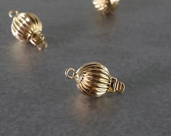 May include: Close-up of several gold-colored, ribbed ball clasps. Each clasp has a small loop on either end for attaching to jewelry. The clasps are on a gray surface, with some in focus and others blurred in the background.