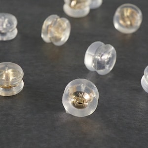 Peut inclure: Gros plan sur dix dos d'oreille en plastique transparent avec un centre en métal doré. Les dos d'oreille sont éparpillés sur une surface grise.