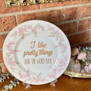 May include: A white ceramic decorative plate with a gold rim and a delicate pink floral pattern around the border. The centre of the plate features gold cursive and block lettering that reads, I like pretty things AND THE WORD FUCK.