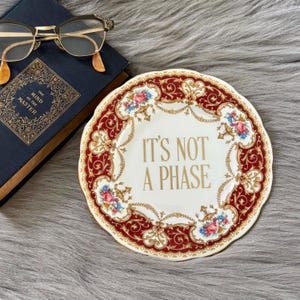 May include: A decorative white plate with a red and gold floral border and the text "IT'S NOT A PHASE" in gold lettering. The plate has a scalloped edge and is decorated with pink and blue flowers. The plate is on a gray surface.