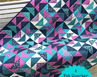 Puede incluir: Un patrón de colcha llamado "Blackberry Twirl" de Jane Michel Quilt Designs. La colcha está hecha con una variedad de telas en tonos de rosa, azul y blanco. La colcha es un patrón amigable con cuartos de tela y mide 122 cm por 152 cm. La colcha se muestra en un banco blanco.