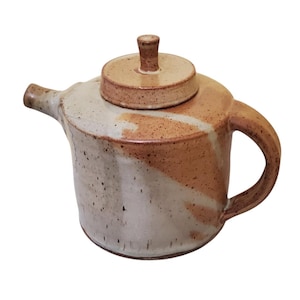 May include: A ceramic teapot with a lid. The teapot is a mix of beige and brown with a handle and spout. The lid has a small knob on top. The teapot is likely handmade and has a rustic appearance.