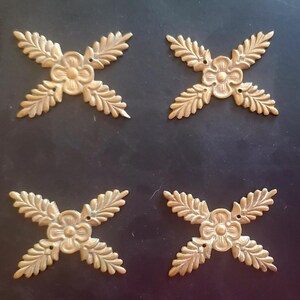 May include: Four antique brass-colored decorative floral ornaments. Each ornament features a central flower with four radiating leaf-like elements. The ornaments are arranged on a dark background.