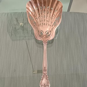 以下が含まれることがあります： 貝殻型のボウルと装飾的なハンドルが付いた銀色のサービングスプーン。 ボウルは繊細な透かし彫りのデザインです。 ハンドルには詳細な彫刻と装飾的な端が付いています。