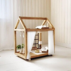 Puede incluir: Una casa de muñecas de madera con forma de casa. El interior presenta muebles en miniatura, incluyendo una cama, un sofá y una lámpara. Cortinas blancas cuelgan a los lados, y un columpio cuelga de la parte superior. La casa de muñecas está sobre ruedas.