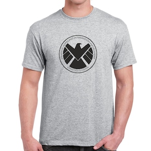 May include: A gray t-shirt with a black logo of the Strategic Homeland Intervention Enforcement and Logistics Division. The logo features a black eagle with a shield and the text "Strategic Homeland Intervention Enforcement Logistics Division".
