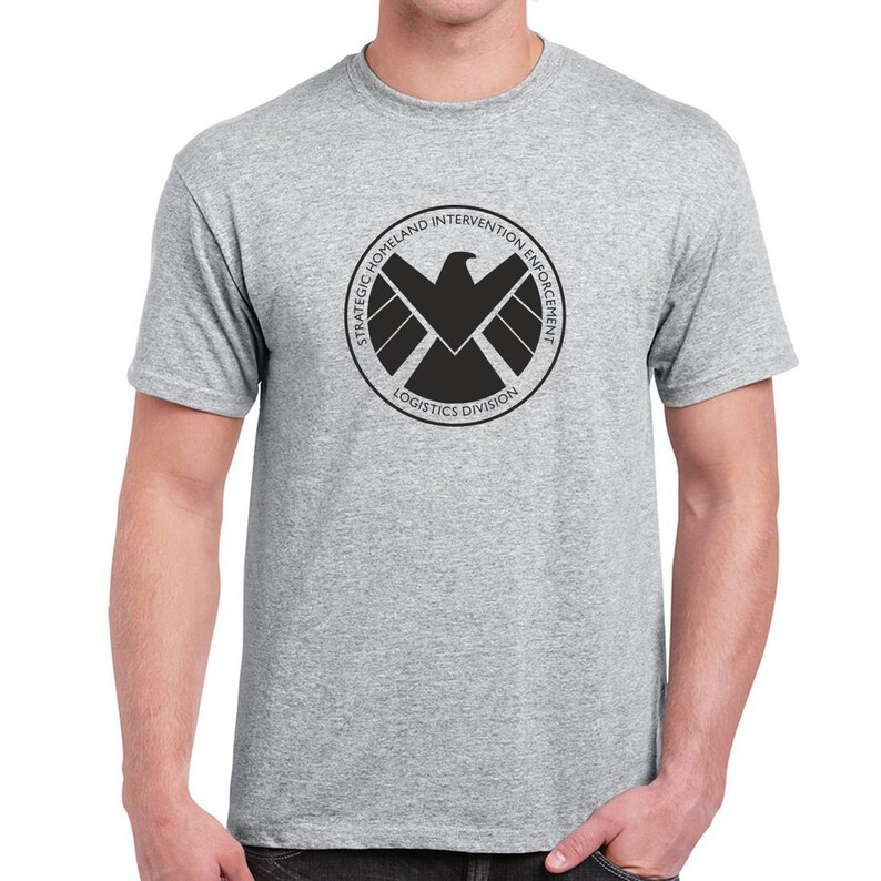 May include: A gray t-shirt with a black logo of the Strategic Homeland Intervention Enforcement and Logistics Division. The logo features a black eagle with a shield and the text "Strategic Homeland Intervention Enforcement Logistics Division".