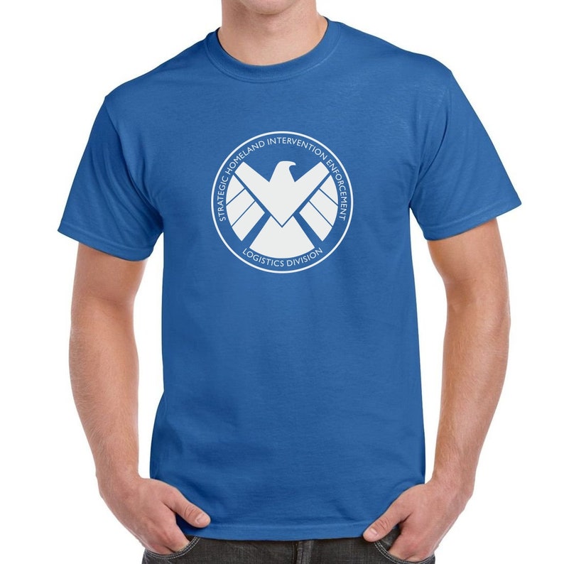 May include: A royal blue t-shirt with a white graphic of the S.H.I.E.L.D. logo. The logo features a white eagle with wings spread and the text "Strategic Homeland Intervention Enforcement Logistics Division".
