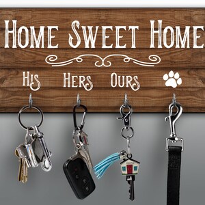 May include: A wooden key holder with the text "HOME SWEET HOME" and "HIS, HERS, OURS". The brown board has a paw print and four metal hooks holding keys and a dog lead. A decorative home key is visible.