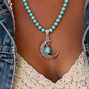 Op de afbeelding: Een ketting met een zilverkleurige halvemaanvormige hanger met gedetailleerd filigraanwerk en een kleine ronde turquoise kraal in het midden. De ketting bestaat uit talloze kleine, ronde, helder turquoise kralen die zijn geregen.