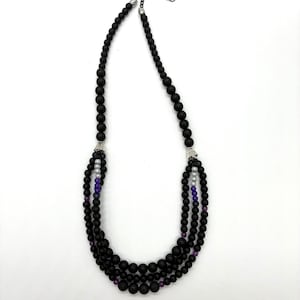 Puede incluir: Un collar de varias hebras con cuentas negras, intercaladas con cuentas moradas y blancas. El collar tiene una cadena y un cierre plateados. Las cuentas están dispuestas en tres capas, creando un efecto de capas. El collar se muestra sobre un fondo blanco.