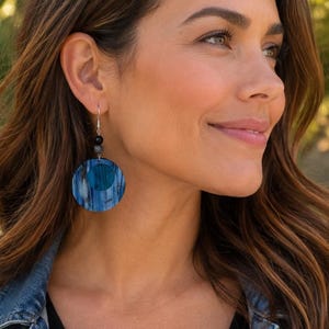 May include: A dangle earring featuring a circular blue and black patterned disc with a solid blue center circle. The earring is suspended from a silver-toned hook by a small black bead and a grey textured bead, creating a layered drop design.