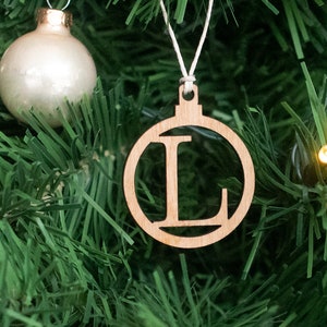May include: A wooden Christmas bauble shaped like a circle with the letter "L" cut out in the centre. The bauble is hanging on a green artificial Christmas tree.