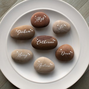 May include: A white plate with seven smooth, grey and brown stones. Each stone has a name written on it in white script with small hearts on either side of the name. The names are: Alyss, Graham, Catherine, Harry, Dena, Andrew, and Jo.