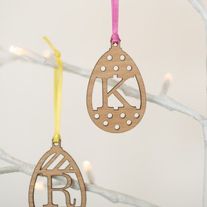 May include: Two wooden Easter egg decorations with ribbon hangers. The egg on the left has a striped design and the letter "R" cut out. The egg on the right has a polka dot design and the letter "K" cut out.