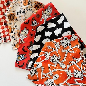 May include: A collection of five triangular fabric bandanas featuring various Halloween-themed patterns including red and white checkered fabric, ghosts with pumpkins, cartoon dogs in costumes, white ghosts on black, and white skeletons on orange.
