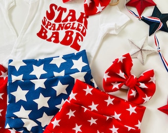 Könnte beinhalten: Ein weißer Strampler mit rotem Text, der "Star Spangled Babe" sagt. Der Strampler wird mit rot-blau gestreiften Shorts und einem rot-weiß gestreiften Stirnband kombiniert.