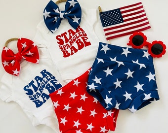 Könnte beinhalten: Zwei weiße Baby-Bodys mit dem Text "Star Spangled Babe" in Rot und Blau. Zwei Paar rote und blaue Baby-Shorts mit Sternenmuster. Eine rot-weiß-blaue Schleife und eine kleine amerikanische Flagge.
