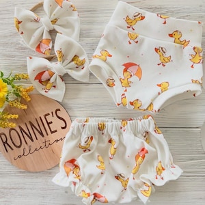 May include: A baby clothing set featuring a white background with a repeating pattern of yellow ducks with umbrellas and hats. The set includes two bows, a pair of shorts, and a pair of ruffled bloomers. A wooden sign with the text "RONNIE'S collection" is also in the image.