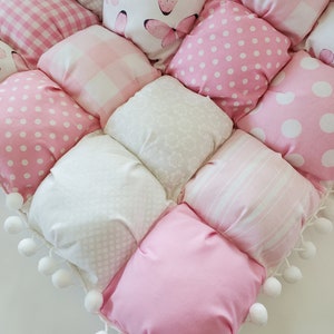 Op de afbeelding: Een handgemaakte, gewatteerde deken met een patchwork-ontwerp van roze en witte vierkanten. De vierkanten tonen verschillende patronen, waaronder stippen, ruitjes, strepen, bloemen en vlinderprints. Witte pompom-bies trimt de deken.