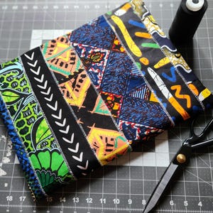 May include: A folded, patchwork fabric item with vibrant patterns in blue, green, yellow, and black. White stitching outlines the fabric sections. A black spool of thread and scissors are visible on a gray cutting mat.