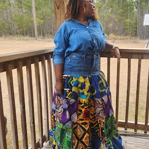 May include: A denim shirt and jeans combination paired with a vibrant, patchwork maxi skirt. The skirt features a mix of colorful African-inspired patterns in blue, yellow, green, and purple. The outfit is worn outdoors on a wooden deck.