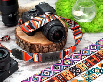 May include: A camera strap with a colorful geometric pattern. The strap is made of woven fabric and has a leather patch with the name "Thomas Murphy" printed on it. The strap is shown attached to a black camera.