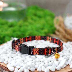 May include: A colorful cat collar with a geometric pattern in shades of orange, red, and black. The collar has a black buckle and a small gold bell.