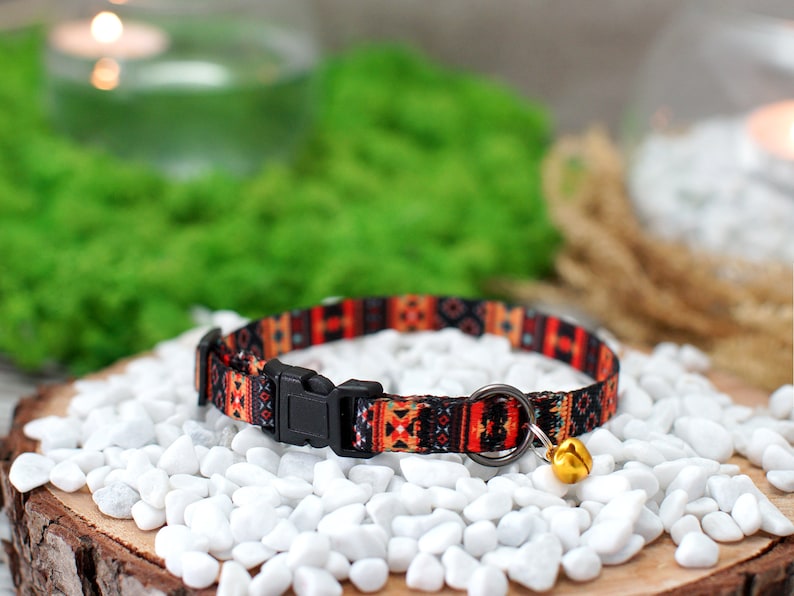 May include: A colorful cat collar with a geometric pattern in shades of orange, red, and black. The collar has a black buckle and a small gold bell.