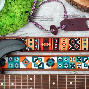May include: A guitar strap with a colorful geometric pattern in shades of orange, blue, and white. The strap is made of a brown leather-like material and has a black adjustable buckle.