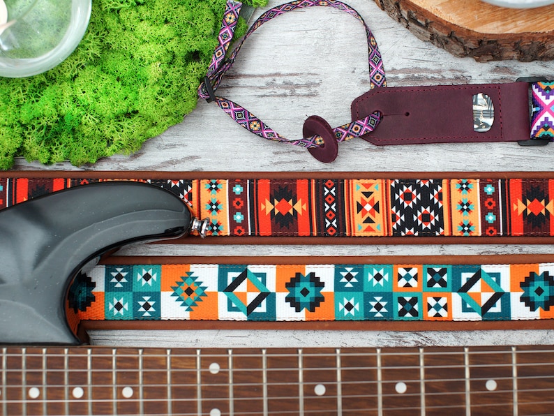 May include: A guitar strap with a colorful geometric pattern in shades of orange, blue, and white. The strap is made of a brown leather-like material and has a black adjustable buckle.