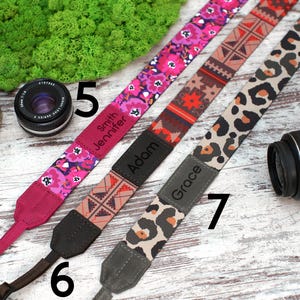 May include: Three camera straps with different patterns and personalised names. The straps are pink with floral print, red and black geometric print, and brown and beige animal print. The names on the straps are "Jennifer Smith", "Adam", and "Grace".
