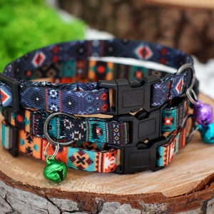May include: Three colourful patterned cat collars with adjustable buckles and small bells. The collars are layered on top of each other, with the top collar being a dark blue with red, white, and black geometric patterns. The middle collar is a bright orange with black, white, and teal geometric patterns. The bottom collar is a teal with black, white, and orange geometric patterns.