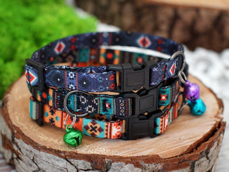 May include: Three colourful patterned cat collars with adjustable buckles and small bells. The collars are layered on top of each other, with the top collar being a dark blue with red, white, and black geometric patterns. The middle collar is a bright orange with black, white, and teal geometric patterns. The bottom collar is a teal with black, white, and orange geometric patterns.