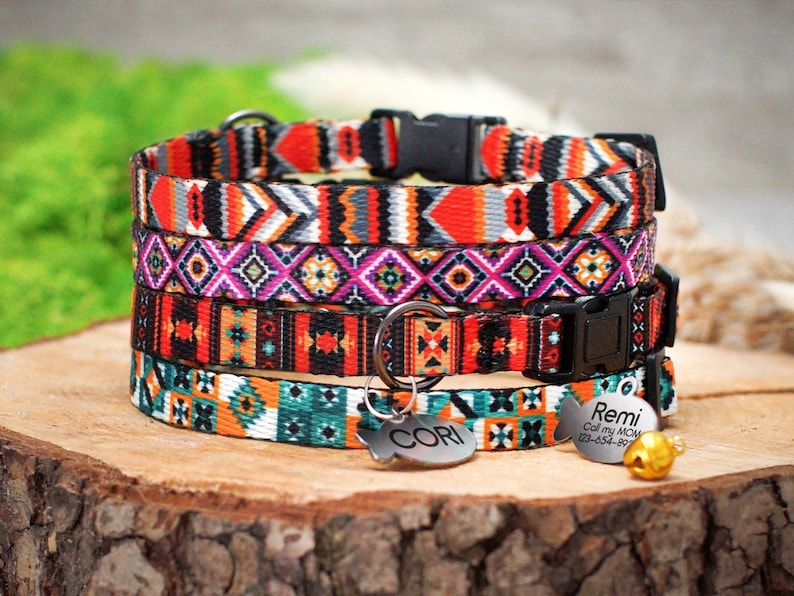 Op de afbeelding: Vier kleurrijke kattenhalsbanden met geometrische patronen in tinten oranje, rood, zwart, wit, teal en roze. De halsbanden hebben zwarte gespen en zijn op elkaar gestapeld. Twee zilveren visvormige labels zijn bevestigd aan de onderste halsband, een met de naam "Cori" en de andere met de naam "Remi" en contactgegevens.