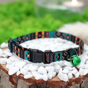 May include: A colorful cat collar with a black buckle and a green bell. The collar has a geometric pattern in shades of blue, orange, and black.