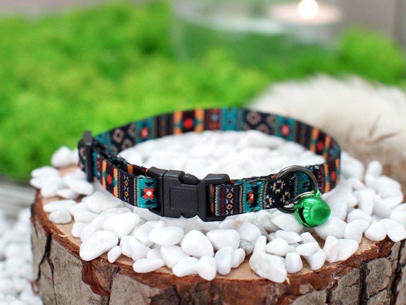 May include: A colorful cat collar with a black buckle and a green bell. The collar has a geometric pattern in shades of blue, orange, and black.